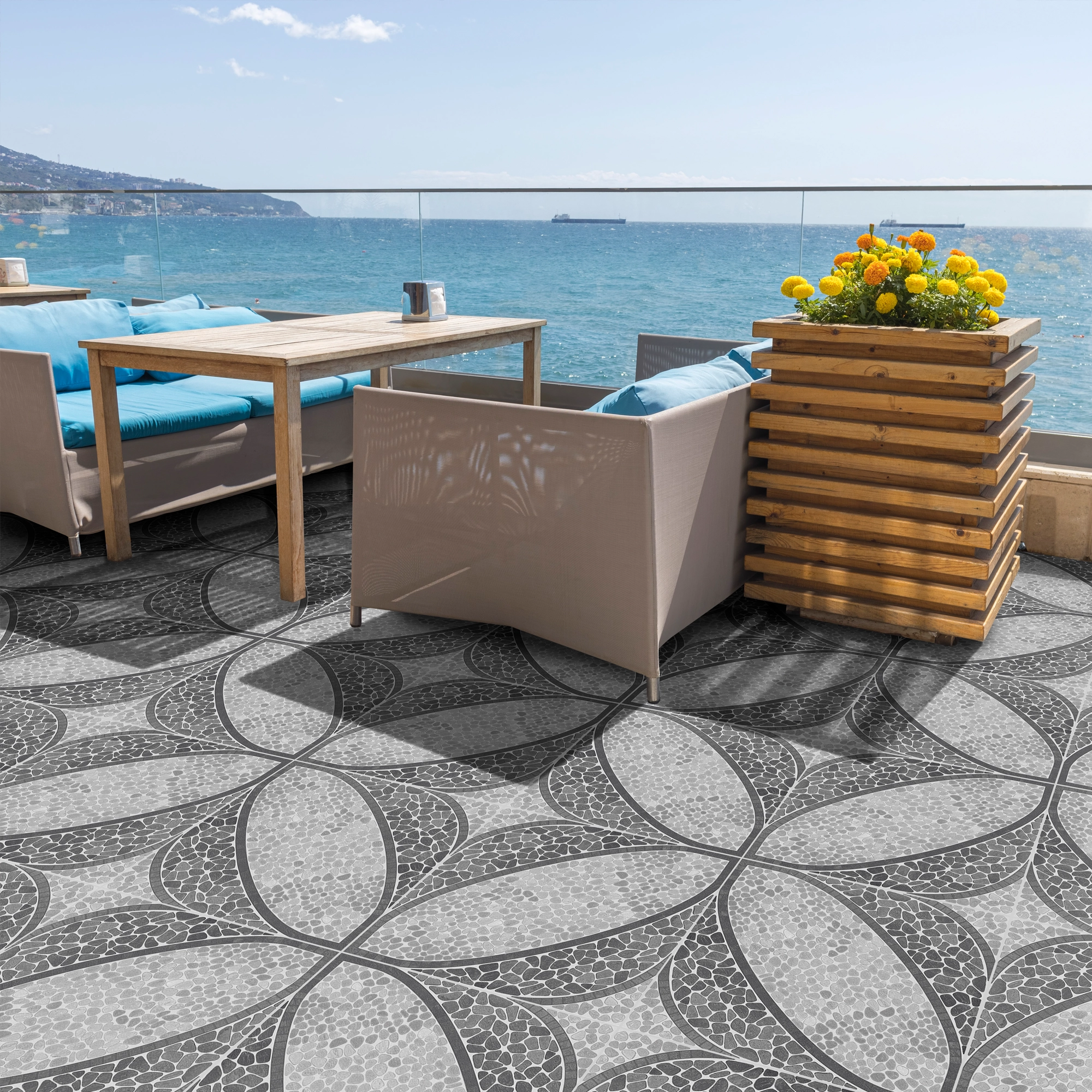 Terraza vista al mar, mesa de madera y asientos con cojines celestes, flores amarilla y piso de cerámica estilo piedra Granara Gris, colección Ecos de cerámicos Gala